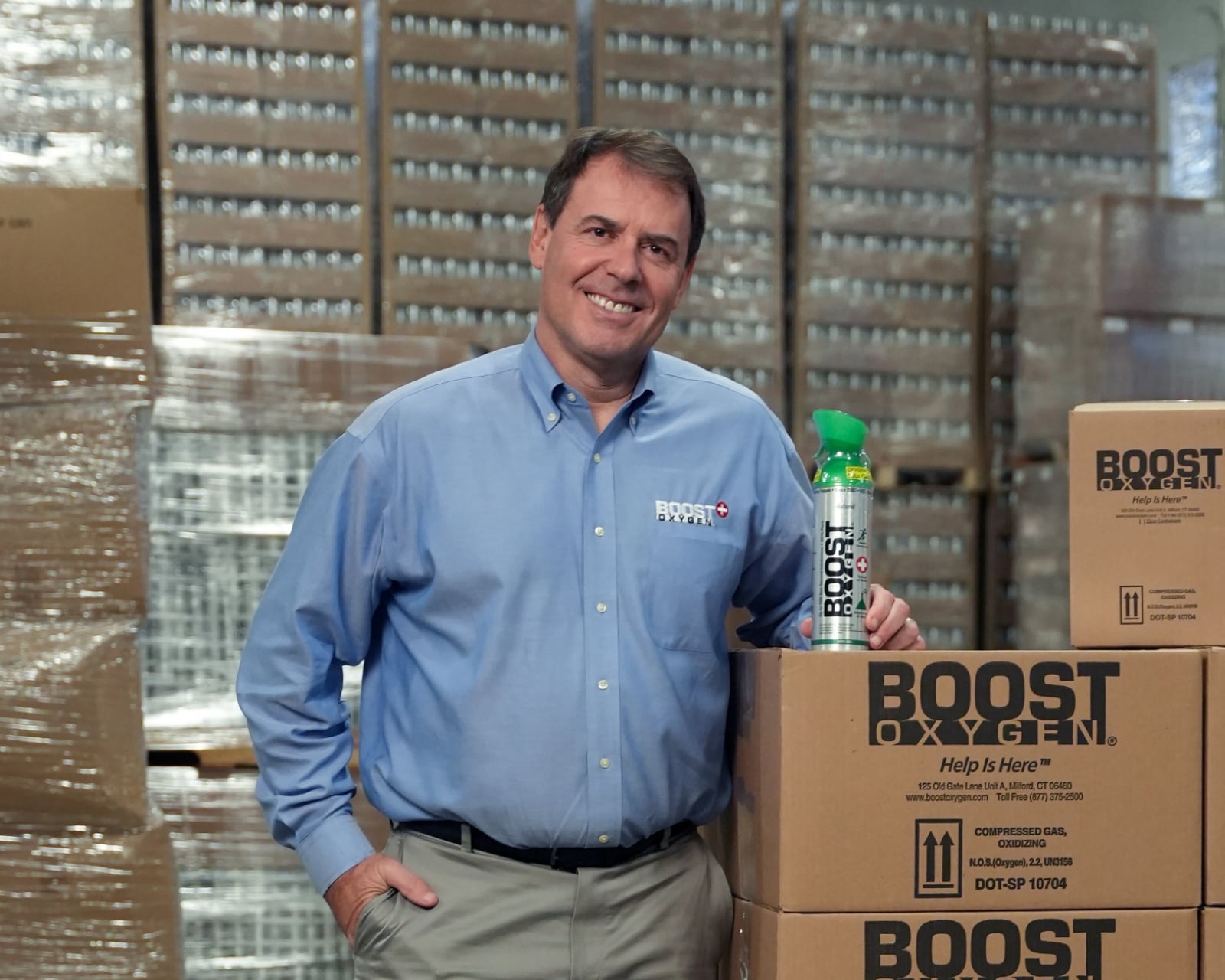 A man in a blue button-down shirt stands smiling with one hand on stacked Boost Oxygen boxes and the other holding a Boost Oxygen canister, highlighting the brand’s ecommerce success amid shelves of boxed goods in the background.